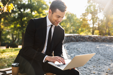 Image of a handsome happy young businessman walking outdoors using laptop computer.の写真素材