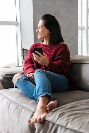 Image of a beautiful woman indoors in home on sofa using mobile phone.の写真素材