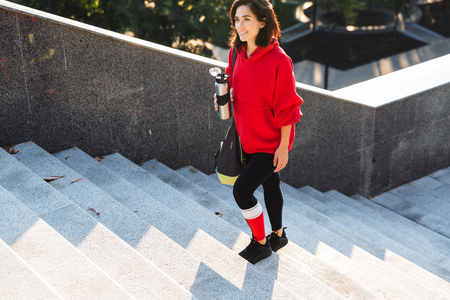 Smiling young sportswoman wearing a hoodie walking outdoors, carrying sports bag, holding coffee cupの写真素材