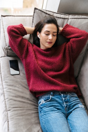 Image of a beautiful woman indoors in home on sofa listening music with earphones.の写真素材