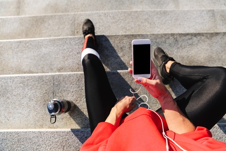 Top view of a young sportswoman holding mobile phone, listening to music with earphones, drinking waterの写真素材