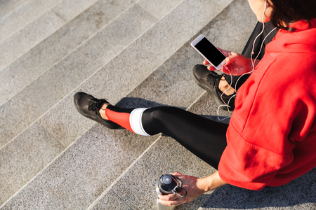 Top view of a young sportswoman holding mobile phone, listening to music with earphones, drinking waterの写真素材