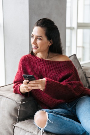 Image of a beautiful woman indoors in home on sofa using mobile phone.の写真素材