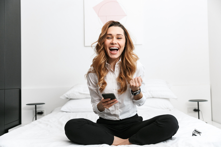 Image of young business woman dressed in formal clothes shirt indoors sitting on bed using mobile phone.の写真素材