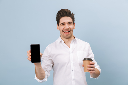 Portrait of a cheerful handsome young man standing isolated over blue background, holding takeaway coffee cup, showing blank screen mobile phoneの写真素材