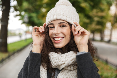 Portrait closeup of happy woman 20s wearing hat and scarf smiling while walking through green parkの写真素材