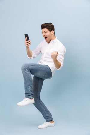 Full length portrait of a cheerful handsome young man standing isolated over blue background, holding mobile phone, celebratingの写真素材