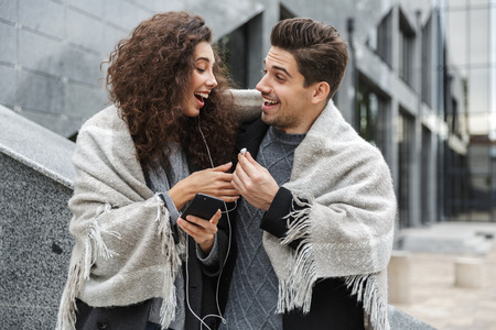 Image of caucasian couple man and woman wearing earphones listening to music together while standing over gray building outdoorの写真素材