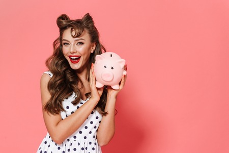 Portrait of a beautiful young pin-up girl wearing dress standing isolated over pink background, showing piggybankの写真素材