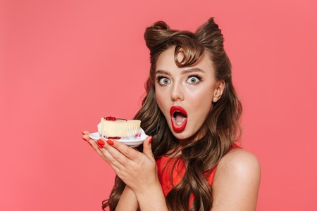 Portrait of a beautiful young pin-up girl wearing dress standing isolated over pink background, eating dessertの写真素材