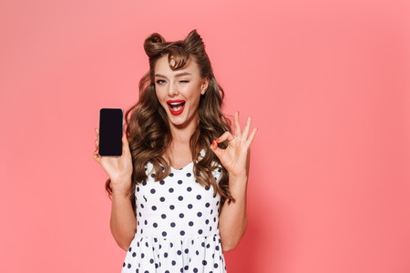 Portrait of a beautiful young pin-up girl wearing dress standing isolated over pink background, showing blank screen mobile phoneの写真素材