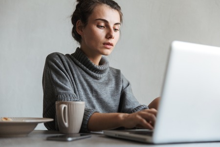 Beautiful young woman having healthy breakfast while sitting on a kitchen, using laptopの写真素材