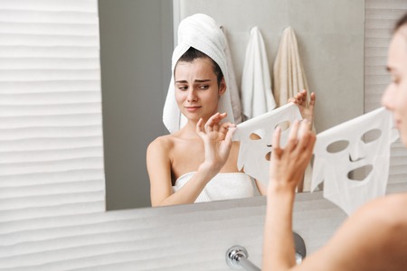 Beautiful young woman with towel on her head standing at the bathroom, trying to apply facial maskの写真素材