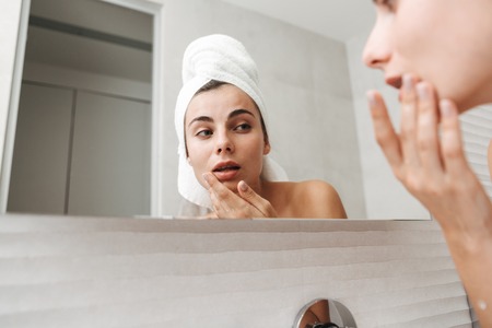 Beautiful young woman with towel on her head standing at the bathroom, examining her face in a...
