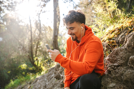Picture of handsome happy young sports fitness man runner outdoors in park listening music with earphones using mobile phone.の写真素材