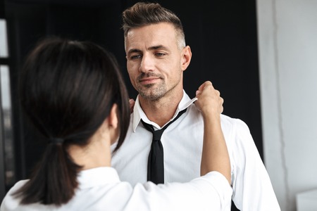 Image of a young woman help to her husband boyfriend with tie formal clothes indoors at home.の写真素材