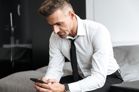 Photo of a business man in bedroom indoors at home sitting on bed using mobile phone.の写真素材