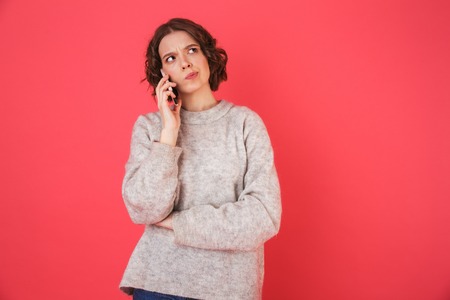 Portrait of a pensive young woman standing isolated over pink background, talking on mobile phoneの写真素材