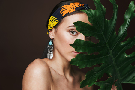 Beauty portrait of a topless young beautiful woman wearing headband and earrings standing isolated over black background, posing with green tropical leafの写真素材