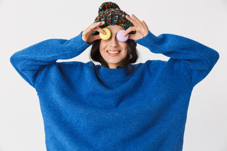 Portrait of optimistic woman 30s wearing winter hat holding macaron cookies while standing isolated...