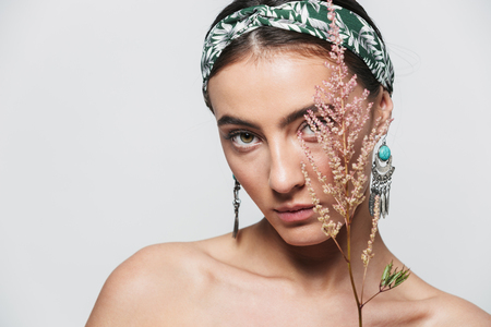 Beauty portrait of a topless young beautiful woman wearing headband and earrings standing isolated over white background, posing with a flowerの写真素材