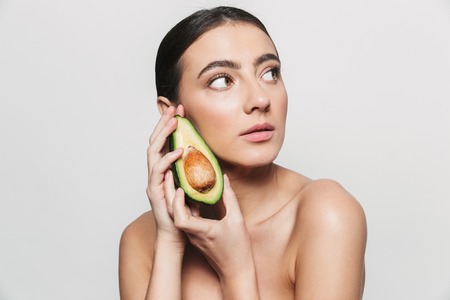 Beauty portrait of a young healthy attractive brunette woman standing isolated over white background, posing with a sliced avocadoの写真素材