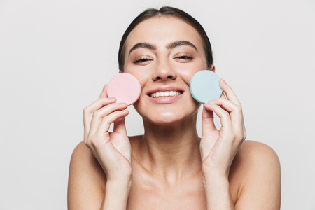 Beauty portrait of a young healthy attractive brunette woman standing isolated over white background, showing makeup spongesの写真素材