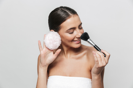 Beauty portrait of a pretty young woman wrapped in towel standing isolated over white background, holding powder puff and a makeup brushの写真素材
