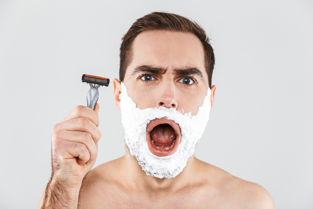 Portrait of a cheerful shirtless bearded man standing isolated over white background, face covered with shaving foam, holding a razorの写真素材