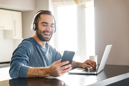 Happy handsome man freelancer sitting at the kitchen table, working on laptop computer, wearing headhonesの写真素材