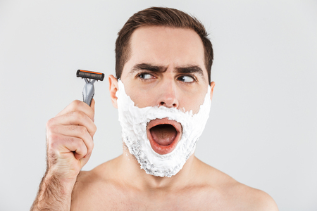 Portrait of a cheerful shirtless bearded man standing isolated over white background, face covered with shaving foam, holding a razorの写真素材