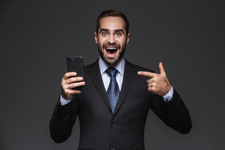 Portrait of a confident handsome businessman wearing a suit standing isolated over black background, using mobile phoneの写真素材