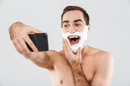 Portrait of a cheerful shirtless bearded man standing isolated over white background, face covered with shaving foam, taking a selfieの写真素材