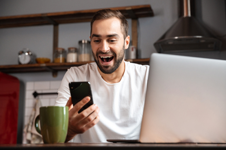 Happy young man using laptop computer while sitting at the kitchen table, holding mobile phoneの写真素材