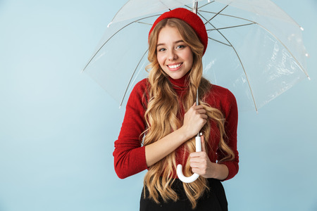 Portrait of cheerful blond woman 20s with long hair standing under umbrella while isolated over blue background in studioの写真素材