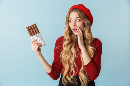 Image of attractive girl 20s wearing red beret eating chocolate bar isolated over blue background in...