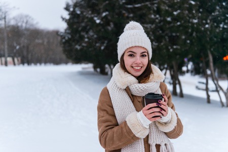 Photo of a pretty young woman in hat and scarf walking outdoors in winter snow drinking coffee.