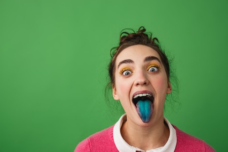 Portrait of a funny young girl standing isolated over green background, showing blue tongue