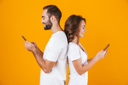 Image of modern couple man and woman smiling while both using mobile phones isolated over yellow backgroundの写真素材