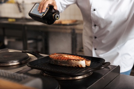 Smiling woman chef cook wearing uniform cooking delicious salmon steak standing at the kitchen, pouring olive oilの写真素材