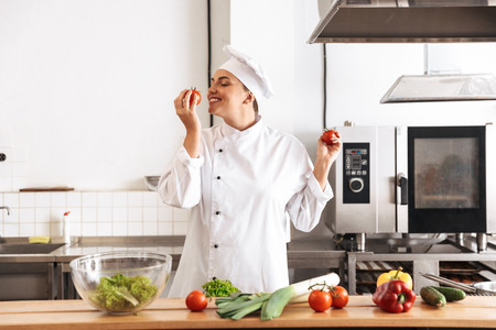 Photo of pleased woman chef wearing white uniform cooking meal with fresh vegetables in kitchen at...