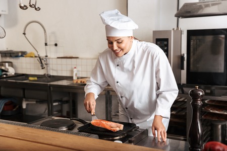 Smiling woman chef cook wearing uniform cooking delicious salmon steak standing at the kitchenの写真素材