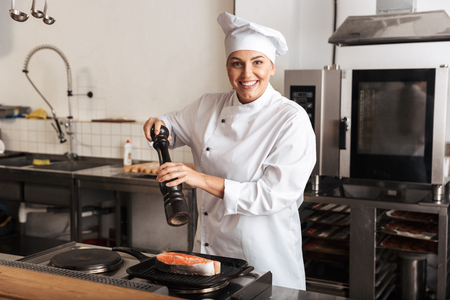 Smiling woman chef cook wearing uniform cooking delicious salmon steak standing at the kitchen