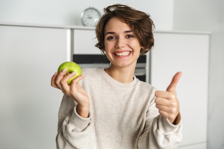 Smiling young woman holding green apple at the kitchenの写真素材