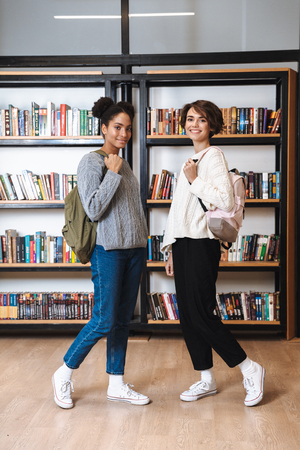 Two cheerful young girls students studying at library, carrying backpacksの写真素材