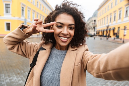 Cheerful young african woman wearing coat walking outdoors, taking a selfieの写真素材