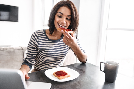 Smiling young african woman having breakfast while sitting at the kitchen with laptop computerの写真素材