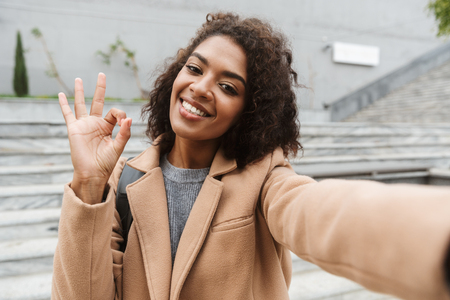 Cheerful young african woman wearing coat walking outdoors, taking a selfieの写真素材