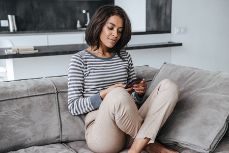 Attractive young afro american woman sitting on a couch at home, using mobile phoneの写真素材