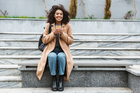 Cheerful young african woman wearing coat sitting outdoors, holding takeaway coffee cupの写真素材